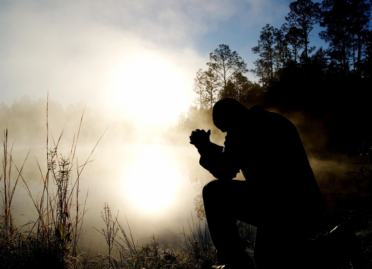 Pessoa em posição de meditação ao ar livre, com as mãos unidas em oração, diante da luz do sol.